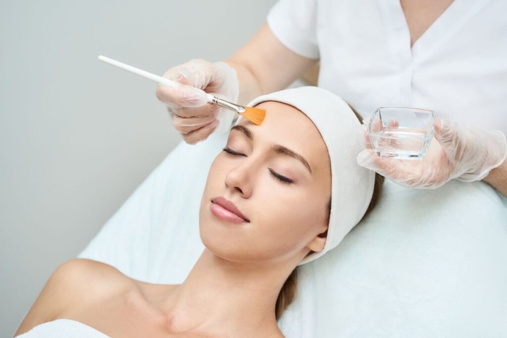 A skincare professional applies a facial treatment to a relaxed client in a spa setting. The client, with a headband and no makeup, lies back on a treatment bed with her eyes closed. The aesthetician, wearing gloves, uses a brush to apply a product from a small glass container. This image highlights the calming atmosphere of a skincare session, showcasing the importance of professional skincare treatments for healthy skin.