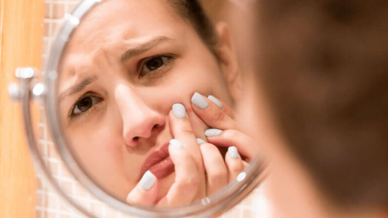 A young woman with light brown hair looks concerned while examining her skin in a bathroom mirror. She is touching a blemish on her face, showcasing a worried expression. The image highlights common skin issues and the emotional impact they can have on individuals. The bathroom setting includes a tiled wall, emphasizing a personal grooming space. This visual representation connects with topics related to skincare, self-care routines, and the challenges of managing acne.