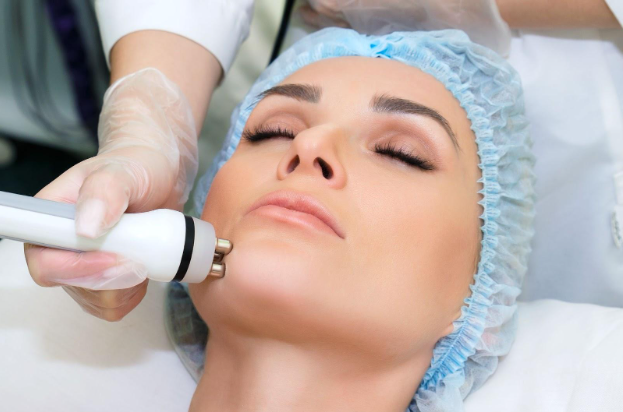 A woman receiving a facial treatment in a spa setting, with a focus on her face. She is lying back, eyes closed, while a technician uses a handheld device for skin therapy. The woman is wearing a hairnet to keep her hair away from her face, and the technician is gloved for hygiene. This image showcases professional skincare techniques aimed at rejuvenating and enhancing facial aesthetics, highlighting the importance of skincare in personal wellness.