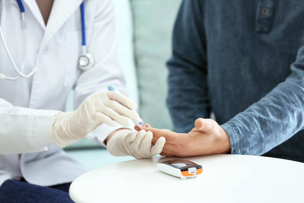 A healthcare professional in a white coat and gloves is performing a blood glucose test on a patient&apos;s fingertip. The patient, wearing a blue long-sleeve shirt, sits at a table with a blood glucose meter positioned nearby. The setting suggests a clinical environment focused on diabetes management and health monitoring. This image emphasizes the importance of regular blood sugar testing for individuals with diabetes.