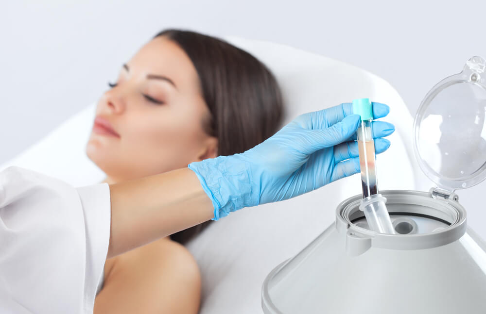 Close-up of a healthcare professional in blue gloves preparing a syringe with a sample, while a relaxed patient lies on a treatment bed. The setting suggests a medical or aesthetic procedure, highlighting the importance of safety and hygiene in patient care. The image conveys a calm environment, emphasizing the professional&apos;s attention to detail in handling medical equipment. Ideal for content related to cosmetic treatments, dermatology, or health and wellness services.