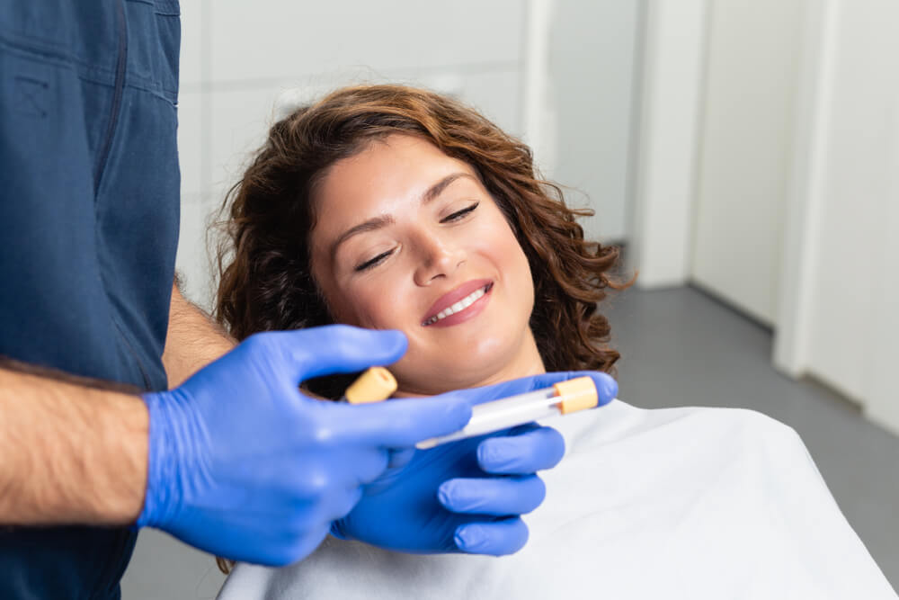 A dental professional in blue gloves prepares a dental instrument while a smiling female patient relaxes in a dental chair. The setting is a modern dental office, highlighting a positive patient experience during a dental procedure. The image conveys professionalism and care, emphasizing the importance of dental health and patient comfort.