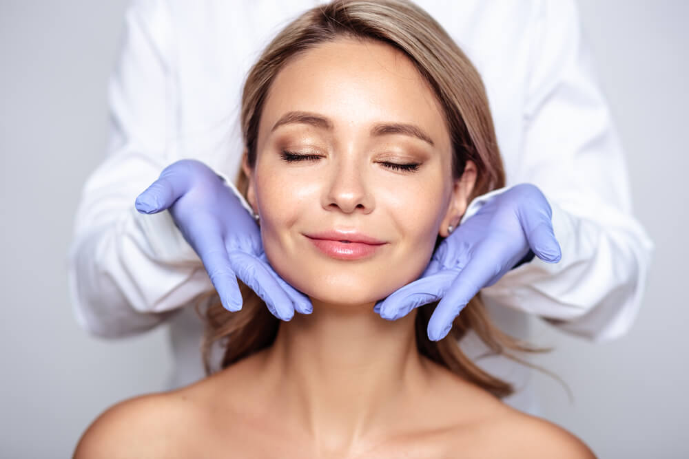 A woman with a serene expression sits with her eyes closed, receiving a facial treatment from a professional in a white coat wearing blue gloves. The setting features a neutral background, emphasizing the focus on the skincare process. This image represents the importance of skincare and beauty treatments, highlighting the expertise of professionals in the field. Ideal for promoting skincare services and wellness practices, it conveys a sense of relaxation and professionalism.