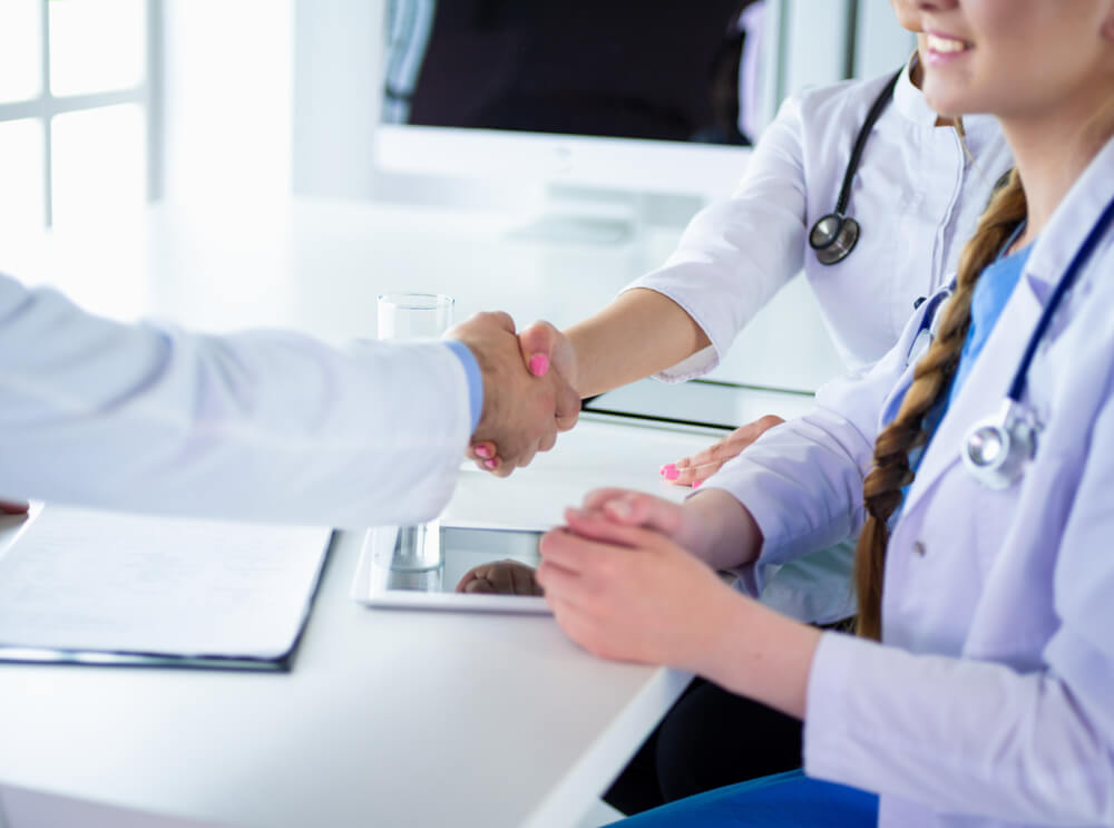 Two healthcare professionals in white lab coats are engaged in a handshake at a modern clinic. One is seated at a table with a tablet, while the other stands and smiles, indicating a positive interaction. A glass of water and a notepad are visible on the table, suggesting a consultation or meeting. The setting is bright and professional, emphasizing a collaborative healthcare environment. This image represents the importance of communication and trust in patient care.