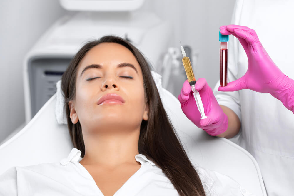 A woman with long hair relaxes in a clinical setting, eyes closed, as a medical professional in pink gloves holds a syringe filled with a golden substance and a test tube containing red liquid. The image illustrates a cosmetic or medical procedure, highlighting the use of injections in skincare treatments. The background features medical equipment, emphasizing a sterile environment. This visual representation is relevant for topics related to beauty treatments, cosmetic procedures, or medical aesthetics.