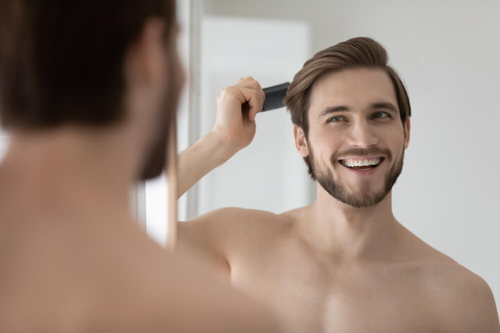 A smiling young man with a beard is grooming his hair with a comb in front of a mirror. He appears happy and relaxed, showcasing a casual morning routine. The bright, neutral-toned bathroom setting emphasizes a clean and fresh atmosphere. This image represents personal grooming, self-care, and confidence, appealing to audiences interested in men&apos;s grooming tips and lifestyle content.