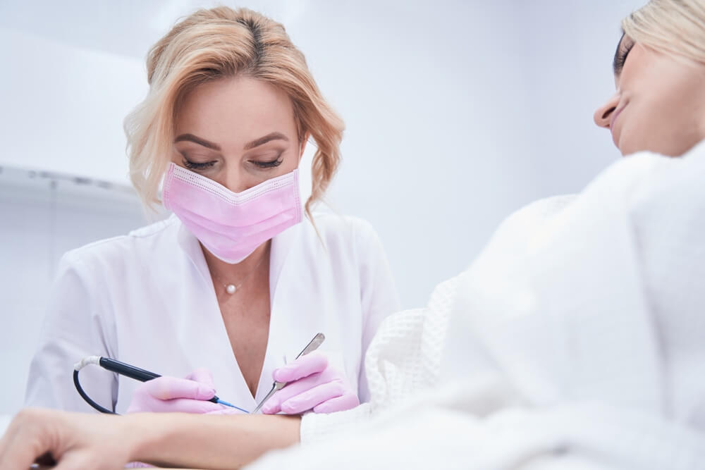 A healthcare professional in a white coat and pink mask is performing a procedure on a patient&apos;s arm in a clinical setting. The practitioner uses a specialized tool while the patient relaxes on a treatment table, indicating a focus on skin care or cosmetic treatment. The sterile environment emphasizes safety and professionalism in the healthcare industry.