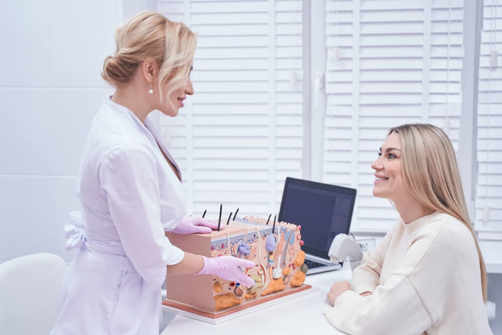 A dermatologist in a white lab coat discusses skin care with a smiling female patient in a modern clinic. The consultation includes a detailed skin model on the table, showcasing various skin treatment tools. A computer is visible in the background, indicating a professional environment focused on skin health and aesthetics. The image highlights a friendly and informative interaction, emphasizing the importance of patient education in dermatology.
