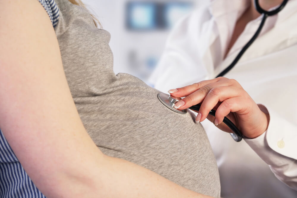 A healthcare professional using a stethoscope to monitor the heartbeat of a pregnant woman. The image captures the close-up of the woman’s rounded belly, emphasizing prenatal care and the bond between mother and healthcare provider. The background suggests a clinical setting, highlighting the importance of regular check-ups during pregnancy for both maternal and fetal health. This visual representation underscores the significance of attentive medical care in ensuring a healthy pregnancy journey.
