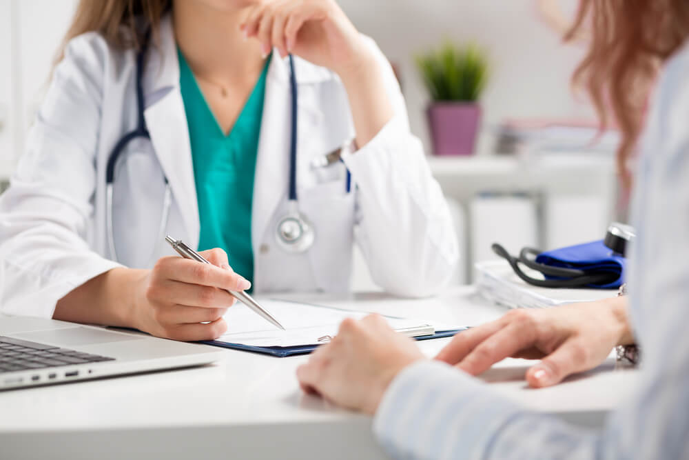 A healthcare professional in a white coat sits at a desk, discussing patient information. The doctor, with a stethoscope around their neck, holds a pen and is reviewing notes while the patient, whose hands are visible, listens attentively. A laptop and medical equipment are on the desk, indicating a clinical environment. This image represents a doctor-patient consultation, emphasizing the importance of communication in healthcare settings.