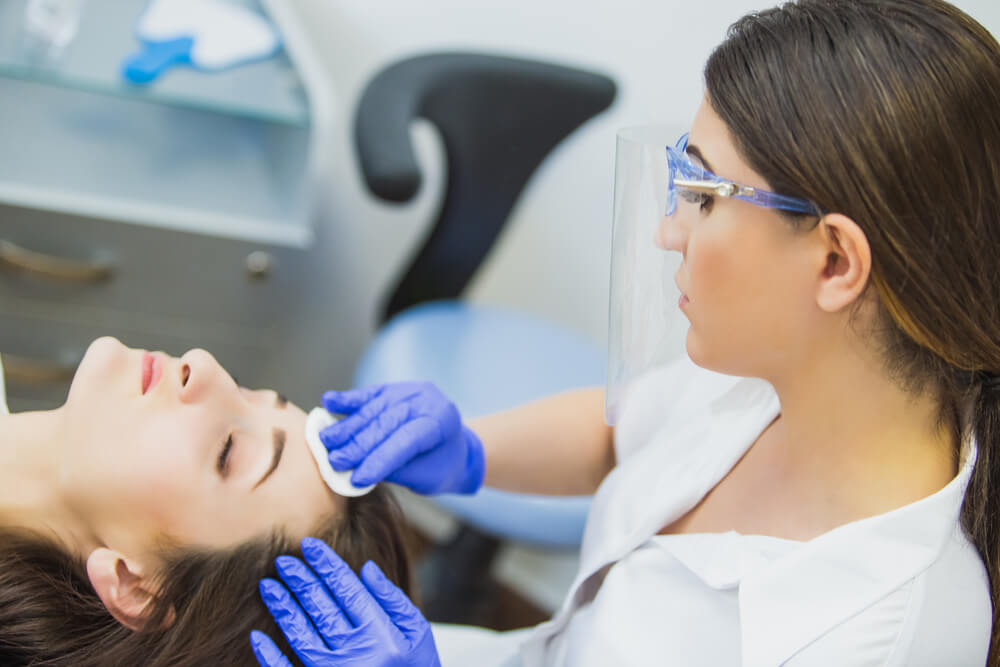 A cosmetologist wearing gloves and a face shield gently cleanses the forehead of a client lying on a treatment bed in a modern skincare clinic. The scene highlights the importance of safety and hygiene in beauty treatments. The background includes a blue chair and skincare tools, emphasizing a professional atmosphere. This image showcases a skincare service focused on client care and comfort, essential for promoting healthy skin.