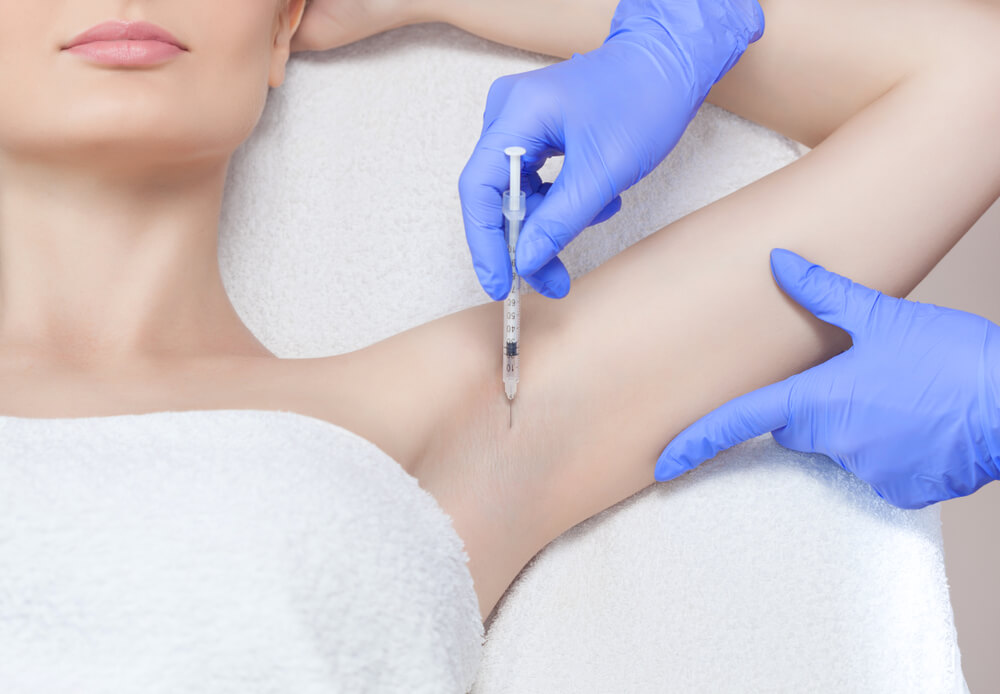 A healthcare professional in blue gloves administers an injection into the underarm area of a patient, who is covered with a white towel. The procedure may be related to cosmetic treatments or medical therapies, highlighting the importance of proper technique and hygiene in clinical settings. This image illustrates the process of injections, emphasizing patient care and safety during medical procedures.