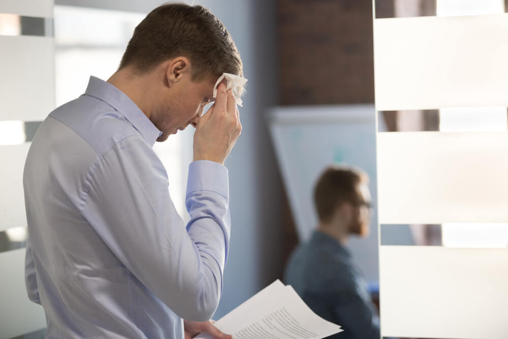 A stressed businessman wipes his forehead with a tissue while holding a document in an office setting. He appears deep in thought, reflecting the pressure of his work environment. In the background, a colleague is seated, engaged in a discussion, highlighting a collaborative workspace. The image conveys themes of workplace stress and the challenges of professional responsibilities.