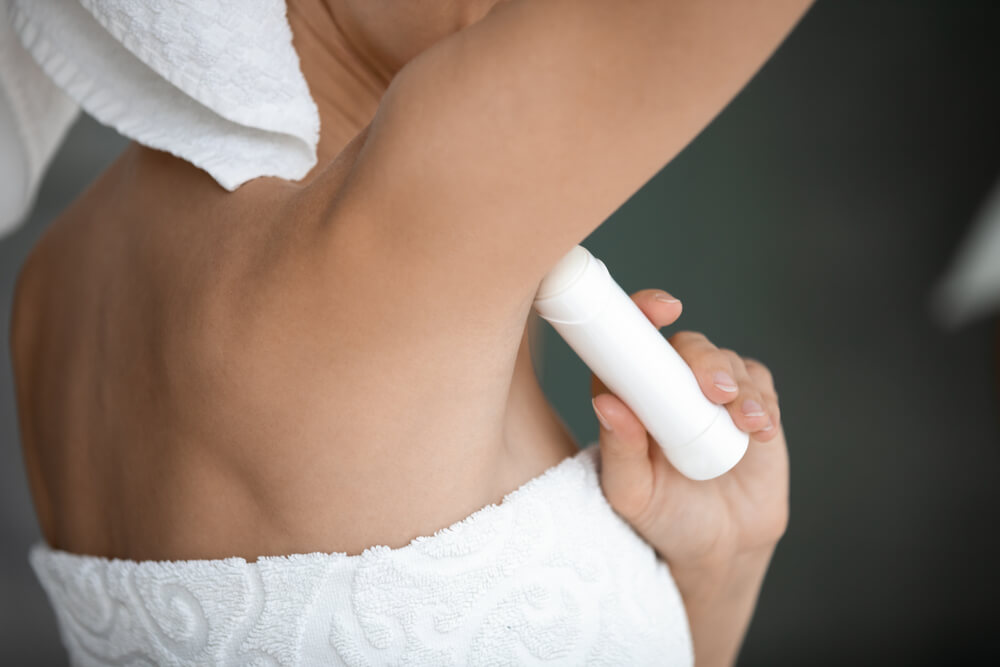 Person applying deodorant to their underarm while wrapped in a white towel, showcasing a personal care routine. The image highlights the importance of hygiene and self-care in daily life. The background is softly blurred, focusing attention on the application process, making it relatable for individuals seeking effective grooming tips. This visual representation promotes awareness of personal grooming products and their use.