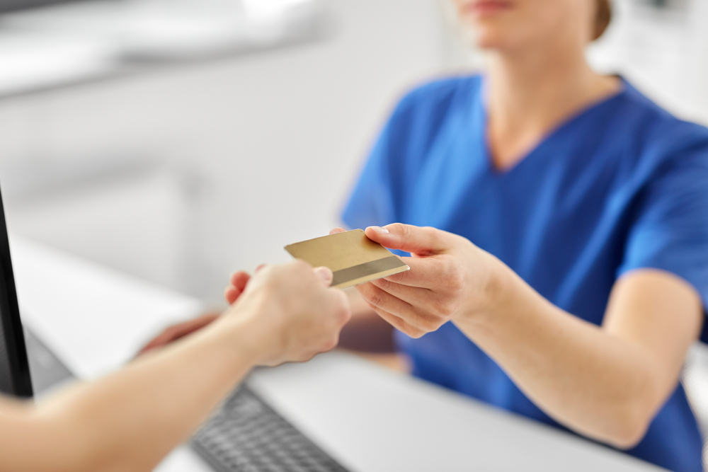 A healthcare professional in blue scrubs hands a gold card to a patient at a reception desk. The setting is a medical office, emphasizing patient care and service. The image highlights the interaction between staff and patients, showcasing the importance of communication in healthcare environments. This visual representation conveys professionalism and a welcoming atmosphere, essential for patient experience in medical facilities.