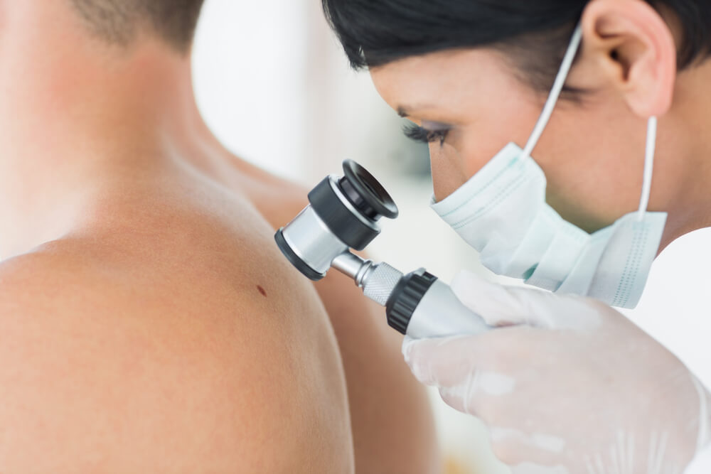 A healthcare professional examines a patient&apos;s back using a dermatoscope, focusing on a skin lesion. The provider wears a mask and gloves, emphasizing safety and hygiene during the dermatological assessment. This image highlights the importance of regular skin checks for early detection of potential skin conditions.