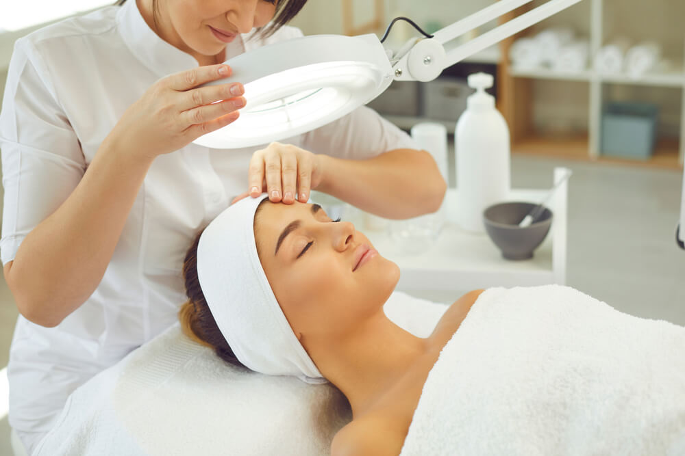 A licensed esthetician performs a facial treatment on a client in a spa setting. The client, lying comfortably on a treatment table, has her eyes closed and a white towel wrapped around her. The esthetician uses a magnifying lamp to examine the client&apos;s skin, ensuring a thorough and precise treatment. In the background, various skincare products are visible on shelves, enhancing the relaxing atmosphere of the spa. This image represents professional skincare services aimed at promoting healthy skin and relaxation.