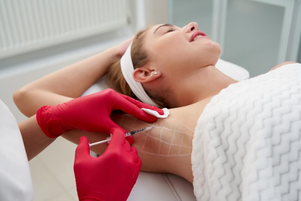A woman receiving a cosmetic treatment on her underarm, with a trained professional in red gloves administering an injection. The setting is a clean and modern clinic, emphasizing safety and professionalism. The woman appears relaxed, lying on a treatment table covered with a soft blanket. This image highlights the process of cosmetic procedures, showcasing the careful attention to hygiene and patient comfort in aesthetic treatments.