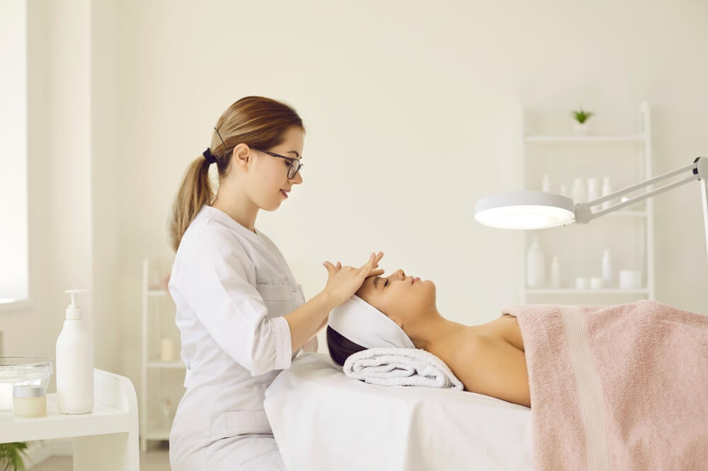 A skincare professional provides a facial treatment to a client in a serene spa environment. The therapist, wearing a white uniform and glasses, gently massages the client&apos;s forehead, who is relaxed and covered with a towel. The treatment area features a well-lit space with skincare products and a magnifying lamp in the background, emphasizing a tranquil atmosphere for beauty and wellness. This image illustrates the importance of self-care and professional skincare services in enhancing skin health.
