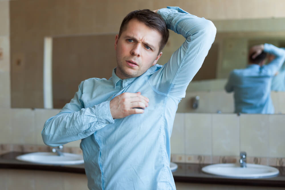 A man in a light blue shirt stands in a bathroom, looking concerned while checking his underarm for sweat. The background features two sinks and a large mirror, reflecting his posture. This image highlights issues related to body odor and personal grooming, emphasizing the importance of confidence and self-care in everyday situations.