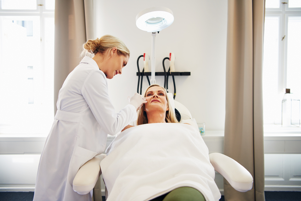 A skincare professional in a white lab coat is performing a facial treatment on a relaxed client lying on a treatment chair. The client, wrapped in a white towel, looks serene under a bright treatment lamp in a well-lit spa environment. The background features soft curtains and skincare tools, creating a calming atmosphere. This image illustrates a professional skincare treatment, highlighting the importance of self-care and wellness in a modern spa setting.