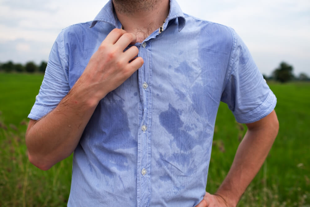 A man stands outdoors in a field, adjusting the collar of his light blue shirt, which is visibly damp with sweat. The background features green grass and a cloudy sky, suggesting warm weather conditions. This image illustrates the effects of heat and physical exertion, highlighting the importance of staying cool and hydrated during outdoor activities.