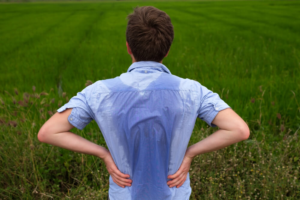 A young man stands with his back to the camera in a lush green field, showing visible sweat on his blue shirt. He has his hands on his hips, suggesting a moment of contemplation or fatigue. The vibrant greenery of the field highlights the outdoor setting, emphasizing the connection to nature and possibly agricultural work. This image captures the essence of hard work and the physical demands of outdoor activities.