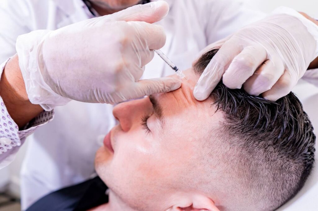 A healthcare professional administers a cosmetic treatment to a male patient, using a syringe to inject a substance into the forehead area. The practitioner wears disposable gloves for hygiene, and the patient appears relaxed with his eyes closed. This image highlights the process of non-surgical aesthetic procedures, often sought for wrinkle reduction and facial rejuvenation.