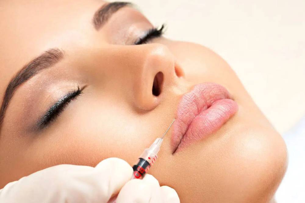 A close-up image of a woman receiving a cosmetic lip filler injection. The practitioner, wearing a glove, holds a syringe near her lips, emphasizing the procedure&apos;s focus on enhancing lip volume and definition. The woman&apos;s eyes are closed, suggesting a calm and relaxed state during the treatment. The background is softly blurred, directing attention to the lip enhancement process, which is popular in cosmetic dermatology for achieving fuller and more youthful lips. This image represents the growing trend in aesthetic treatments aimed at improving facial features.