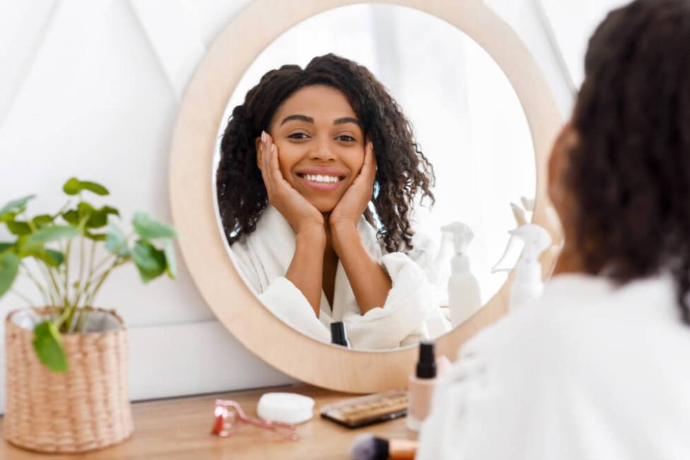 Smiling woman with curly hair in a white robe, sitting in front of a round mirror, showcasing a skincare routine. She has her hands on her face, exuding a sense of joy and self-care. A small potted plant and various skincare products are visible on a wooden vanity, creating a serene and inviting atmosphere. This image highlights themes of beauty, wellness, and personal care, making it ideal for content related to skincare tips, beauty routines, or self-care practices.