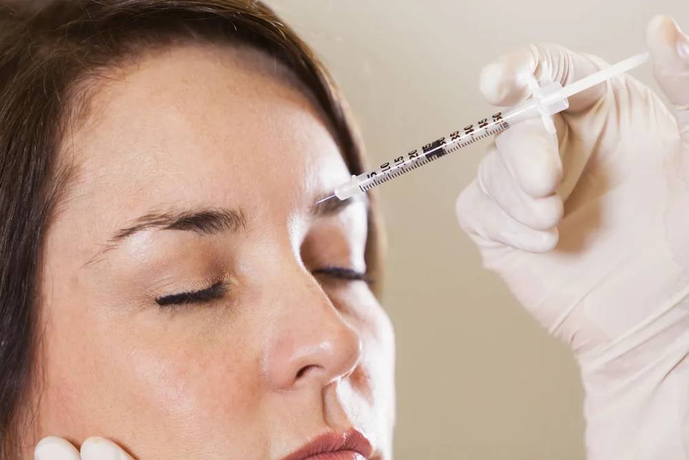 A healthcare professional administers an injectable treatment to a woman’s forehead. The woman has her eyes closed, indicating a calm demeanor during the procedure. The professional is wearing gloves for safety and hygiene. This image represents cosmetic procedures aimed at reducing wrinkles or enhancing facial features, highlighting the importance of professional care in aesthetic treatments.