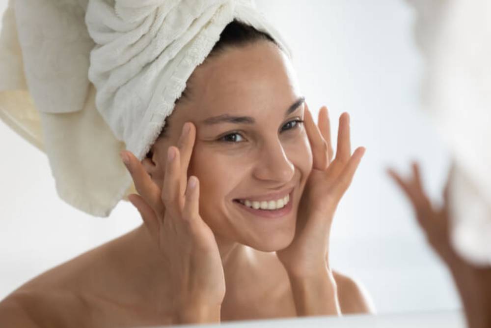 A woman with a towel wrapped around her hair smiles while applying skincare products to her face in front of a mirror. The image captures a moment of self-care and beauty routine, emphasizing the importance of skincare and wellness. The bright, soft lighting and neutral background create a calming atmosphere, highlighting the woman&apos;s glowing skin and positive expression. This image represents a healthy lifestyle and the practice of daily skincare rituals.