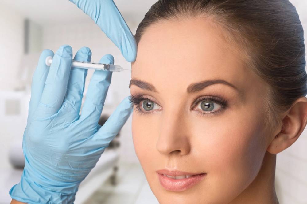 A healthcare professional wearing blue gloves administers a cosmetic injection to a woman's forehead in a clinical setting. The woman has a smooth complexion and is looking directly at the camera, showcasing her natural beauty. The background is softly blurred, emphasizing the procedure. This image highlights the growing trend of cosmetic treatments aimed at enhancing facial aesthetics.
