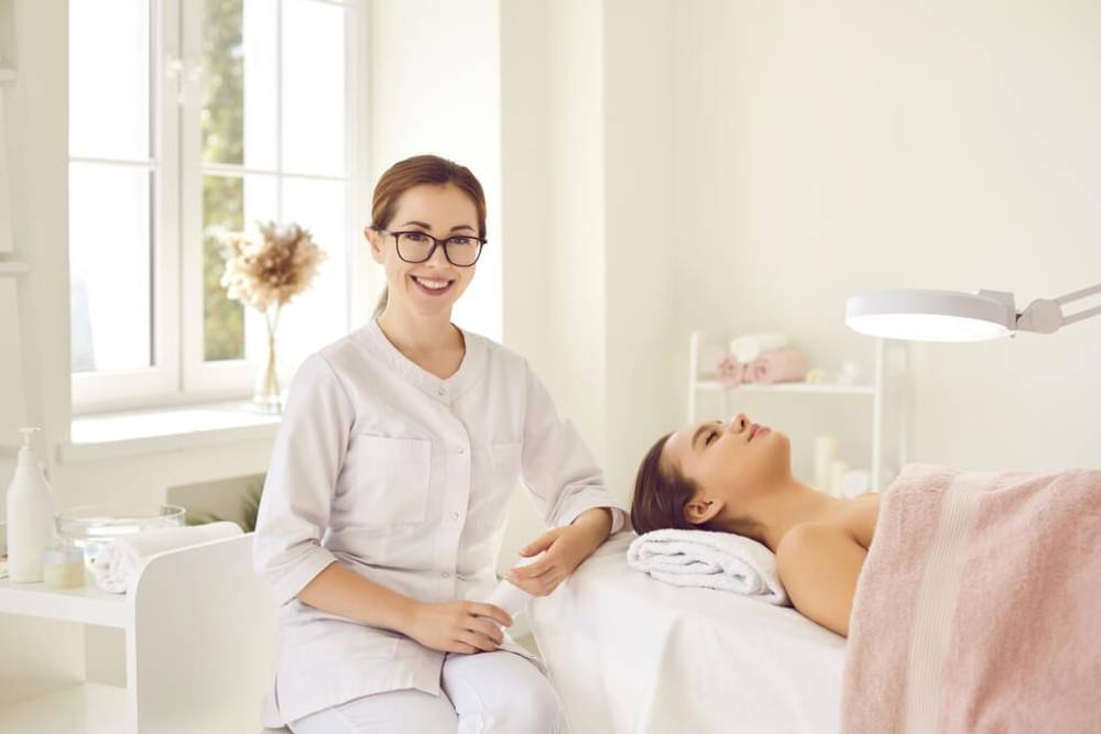 Aesthetician in a spa setting smiles while seated next to a client receiving a facial treatment. The room features natural light from large windows, soft decor, and professional skincare products on display. The client relaxes on a treatment table, covered with a towel, under a magnifying lamp. This image captures a serene atmosphere, highlighting the importance of self-care and professional skincare services. Ideal for promoting wellness, beauty treatments, and spa services.