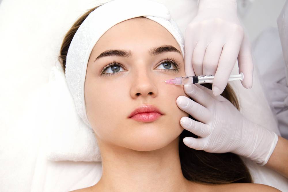 Aesthetic treatment session featuring a young woman receiving an injectable procedure. The practitioner, wearing white gloves, carefully administers a syringe to the woman&apos;s face, focusing on her cheek area. The patient has a headband on to keep her hair away from her face and appears relaxed while lying on a treatment bed. This image illustrates the process of cosmetic enhancements, highlighting the growing popularity of non-surgical beauty treatments.