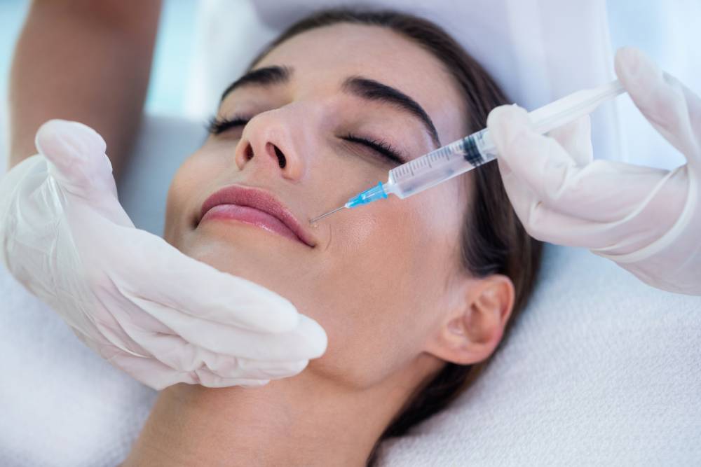A healthcare professional administers a cosmetic injection to a woman&apos;s lips in a clinical setting. The woman appears relaxed as she receives the treatment, which is commonly used for lip enhancement and facial rejuvenation. The practitioner, wearing white gloves, carefully holds the woman&apos;s chin while preparing to inject. This image highlights the growing trend of non-surgical cosmetic procedures aimed at enhancing natural beauty.