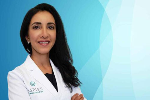 Female dermatologist in a white lab coat stands confidently with arms crossed, smiling against a light blue gradient background. The lab coat features the logo of Aspire Dermatology, indicating her professional affiliation. This image conveys professionalism and expertise in dermatological care, suitable for a medical practice website or promotional materials.