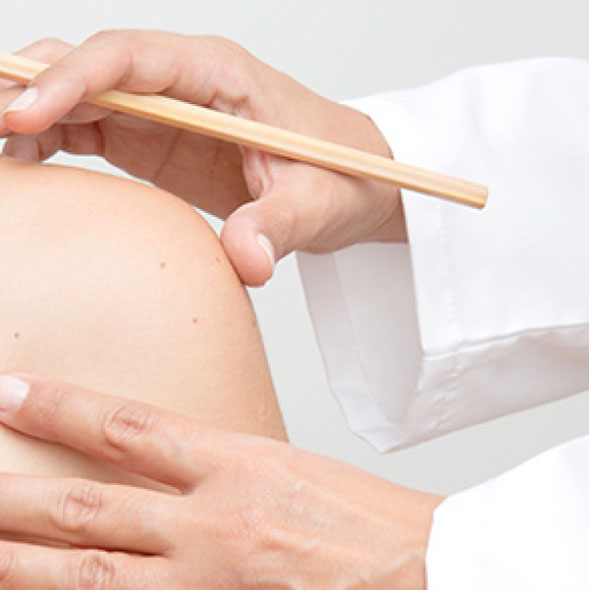 A healthcare professional uses a wooden tool to perform a procedure on a patient&apos;s shoulder. The image highlights the careful technique involved in patient care, emphasizing the importance of precision in therapeutic practices. The setting is clinical, with a focus on the interaction between the practitioner and the patient, showcasing a moment of attentive care.