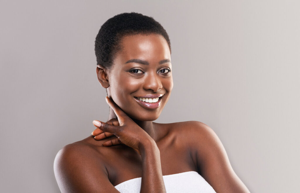 Smiling woman with short hair and glowing skin, wearing a white towel. She is posing against a soft gray background, with her hand gently touching her neck. The image conveys confidence and beauty, ideal for beauty and skincare content.