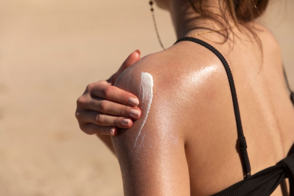 A close-up image of a person applying sunscreen to their shoulder at the beach. The skin appears sun-kissed and glistening, indicating sun exposure. The individual is wearing a black swimsuit top, and the background shows a sandy beach environment. This image emphasizes the importance of sun protection and skincare while enjoying outdoor activities.