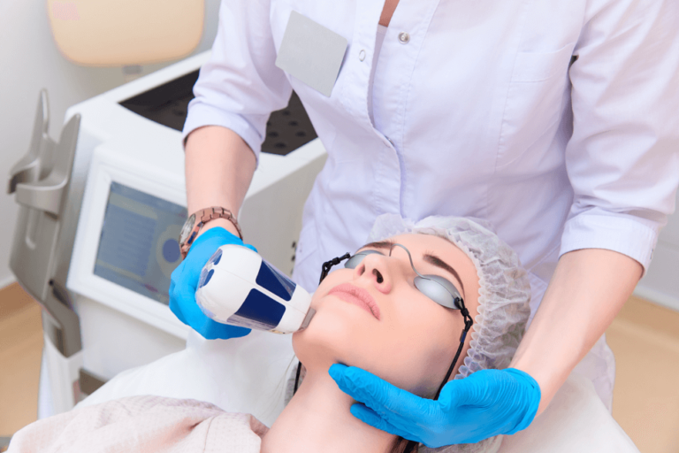 A skincare professional in blue gloves performs a facial treatment on a female client in a clinical setting. The client is lying on a treatment bed, wearing protective eyewear and a hair covering, while the practitioner uses a handheld device on her chin. The background features a modern aesthetic machine, indicating a focus on advanced skincare technology. This image represents the growing trend of professional skincare treatments aimed at enhancing beauty and promoting skin health.