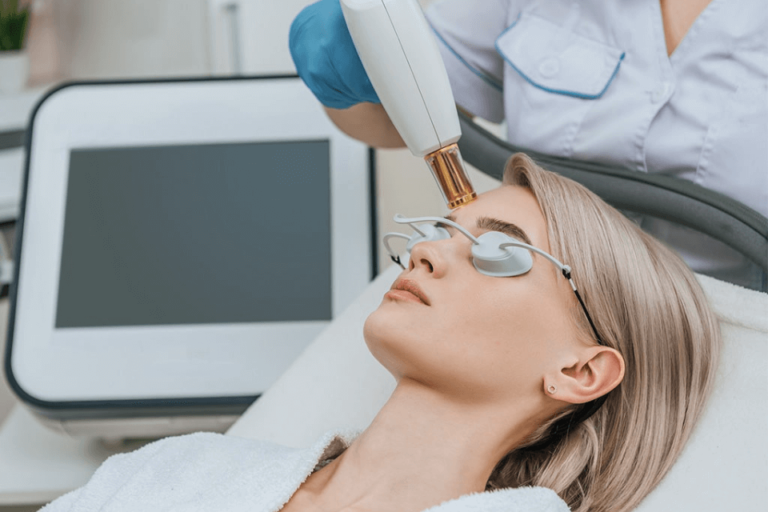 A woman receiving a facial treatment in a skincare clinic, wearing protective eyewear while a technician uses a handheld laser device. The setting features a modern aesthetic with a medical device in the background, highlighting a professional environment focused on skincare and beauty treatments. The woman appears relaxed, indicating a comfortable experience during the procedure. This image illustrates advanced skincare technology and the importance of safety measures in cosmetic treatments.