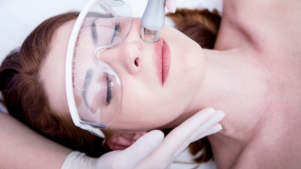 A woman receiving a facial treatment lies on a treatment bed, wearing protective goggles. A licensed aesthetician, gloved and focused, uses a handheld device to perform a skin procedure on her face. The setting is a clean and professional skincare clinic, emphasizing safety and care in cosmetic treatments. This image highlights advanced skincare techniques and the importance of professional guidance in personal beauty routines.