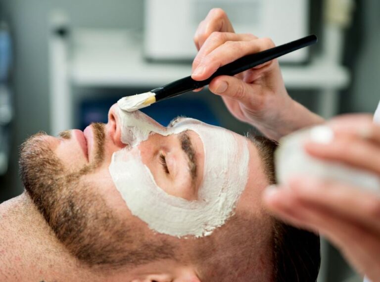 A man receiving a facial treatment at a spa, with a white mask applied to his face. A beautician is gently applying the mask using a brush, while holding a small container of the product in their other hand. The background features spa equipment, creating a relaxing atmosphere. This image highlights skincare and grooming services, emphasizing self-care and wellness.