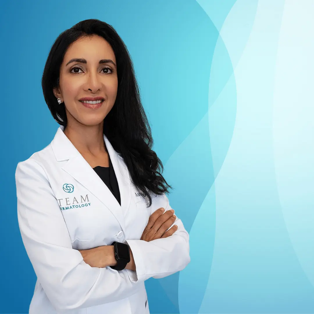 Professional dermatologist wearing a white coat with the logo of "TEAM Dermatology," standing confidently with arms crossed against a light blue gradient background. The image conveys expertise in skincare and dermatological treatments, aimed at building trust with potential patients.