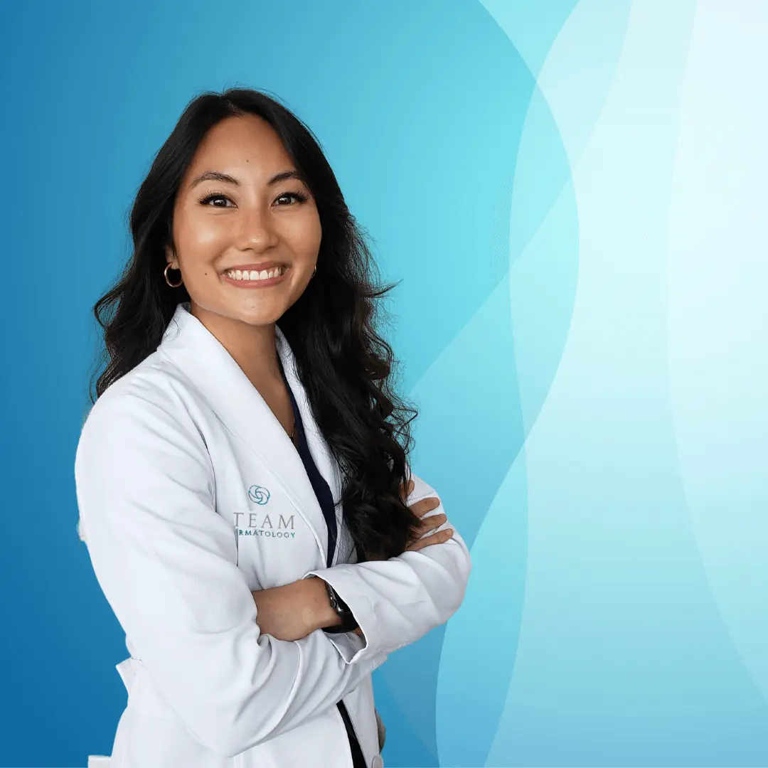 Portrait of a smiling female dermatologist wearing a white lab coat, standing confidently with arms crossed against a soft blue background. The image reflects professionalism and expertise in dermatology, highlighting the importance of skin health and care. Ideal for promoting dermatological services and building trust with potential patients.