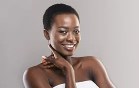 Smiling woman with short hair, dressed in a strapless top, poses confidently against a neutral gray background. Her hand rests gently on her neck, showcasing a warm and inviting expression. The image highlights her radiant skin and bright smile, emphasizing beauty and self-confidence. Ideal for topics related to beauty, fashion, or self-empowerment.