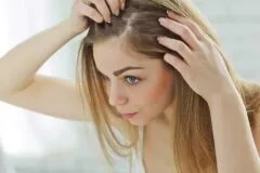 A young woman examines her scalp and hairline in a well-lit bathroom. She has long, straight hair and is using her fingers to part her hair, likely checking for hair loss or scalp health. The image highlights concerns about hair thinning and personal grooming, emphasizing the importance of hair care and self-examination.