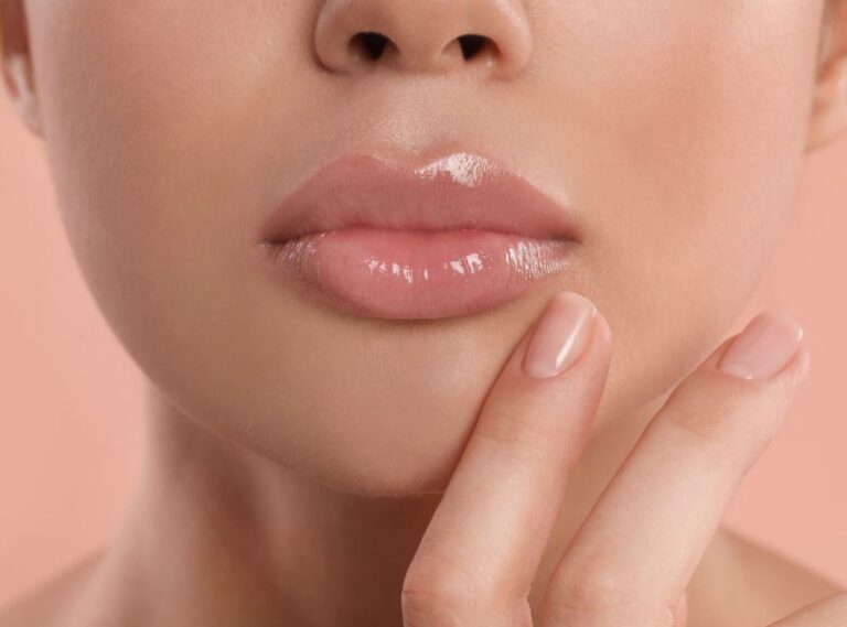 Close-up of a woman&apos;s face highlighting her glossy, full lips and natural makeup. She gently touches her chin with one hand, showcasing a soft, polished manicure. The background features a warm, peach hue, enhancing the overall aesthetic. This image conveys beauty and skincare, suitable for topics related to lip care, makeup techniques, or beauty trends.