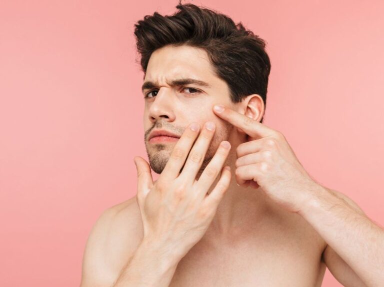 A young man with short dark hair is examining his face, specifically focusing on a blemish on his cheek. He looks concerned as he gently points to the area with one hand while using the other hand to support his chin. The background is a soft pink color, creating a contrasting and visually appealing scene. This image captures a common moment of self-care and concern about skin health, highlighting the importance of skincare routines.